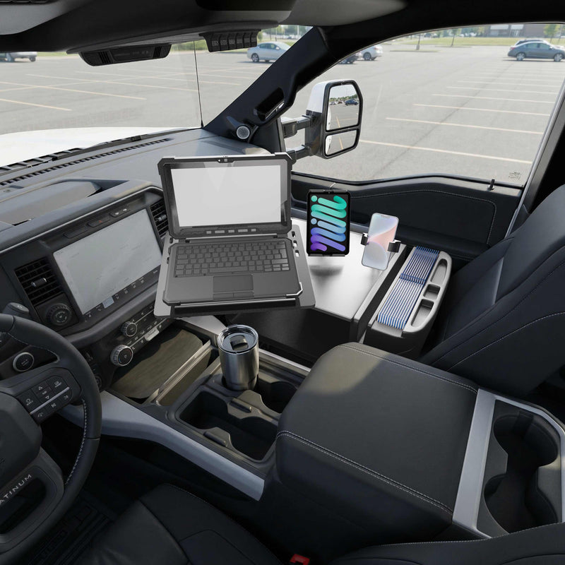A laptop and tablet—secured in the Slim-Grip® Ultra Pro and RoadVise® Ultra Pro Drill-Base Mount—along with wireless earbuds, a travel mug, and neatly organized trays are arranged on the AutoExec Reach Front Seat Car Desk, creating a mobile workstation inside a parked car with an empty parking lot visible through the windows.
