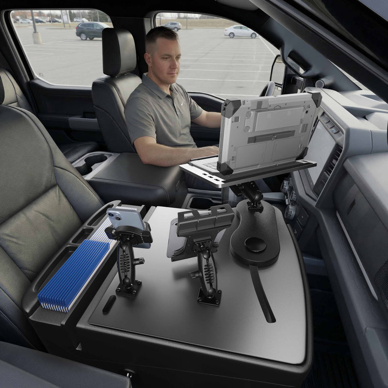 A man sits in the driver’s seat of a parked vehicle, working on an AutoExec Reach Front Seat Car Desk equipped with a Slim-Grip® Ultra Pro and RoadVise® Ultra Pro Drill-Base Mount. Additional device mounts, including a tablet holder, are installed as part of this mobile workstation setup in the front passenger area.