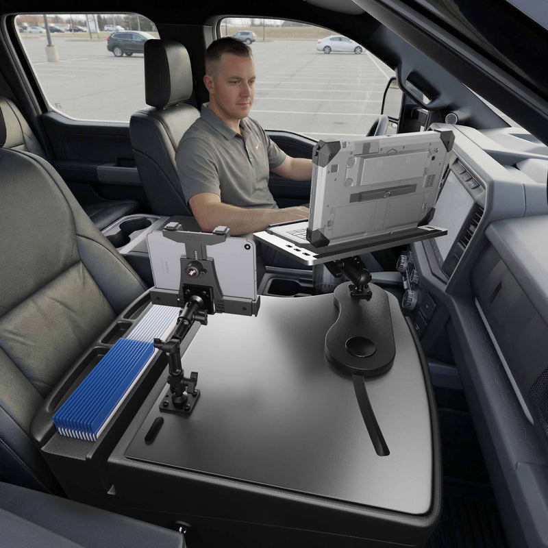 A person sits in the driver’s seat of a parked vehicle, working at an AutoExec Reach Front Seat Car Desk with a LockVise™ Locking Tablet Multi-Angle Mount installed in the front passenger area, which holds a laptop, a securely mounted tablet, and a blue-and-white document organizer.