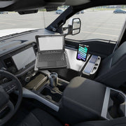 An AutoExec Reach Front Seat Car Desk with Slim-Grip® Ultra Pro Multi-Angle Mount by Arkon Mounts holds a laptop and smartphone inside the front seat area of a parked vehicle. The dashboard, steering wheel, and a cup holder with a metal tumbler are also visible.