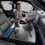 A person sits in the driver’s seat of a parked vehicle, using the AutoExec Reach Front Seat Car Desk with Slim-Grip® Ultra Pro Multi-Angle Mount by Arkon Mounts, positioned over the center console as a mobile workstation.