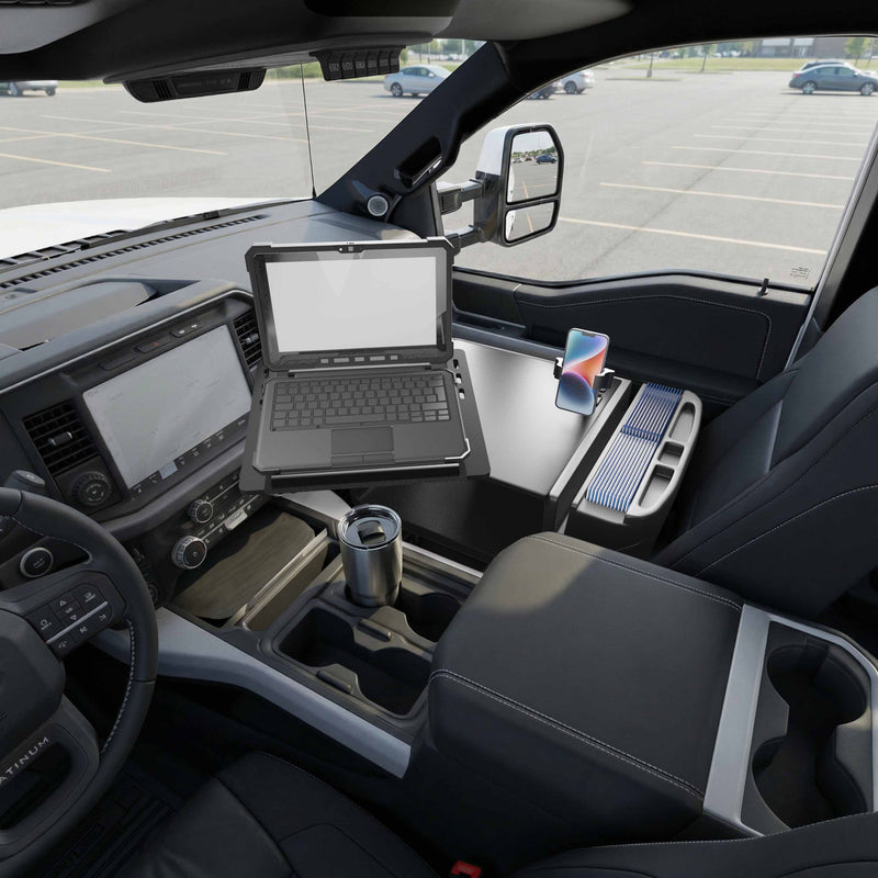 An AutoExec Reach Front Seat Car Desk with RoadVise® Ultra Pro Drill-Base Mount is set up inside a parked truck, supporting a rugged laptop and holding a smartphone securely in its mount beside a coffee tumbler. The modern dashboard with infotainment screen and the view of the parking lot through the window create an efficient mobile workstation on the go.