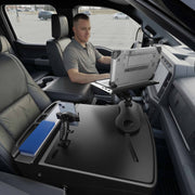 A person sits in a parked vehicle using the AutoExec Reach Front Seat Car Desk with RoadVise® Ultra Pro Drill-Base Mount on the front passenger seat as a mobile workstation, with a parking lot visible outside the window.