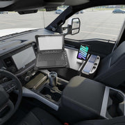 Inside a parked vehicle, an Arkon Mounts AutoExec Reach Front Seat Car Desk with Slim-Grip® Ultra Pro Drill-Base Mount securely holds a rugged laptop and tablet, creating an efficient mobile workspace. A stainless steel travel mug sits in the cup holder while the empty parking lot is visible through the windows, illustrating the practicality of this versatile car desk and tablet holder setup.