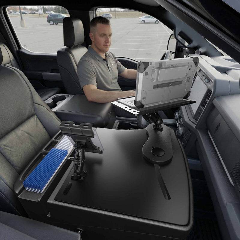 A man sits in the driver’s seat of a parked vehicle, working on his laptop mounted to the AutoExec Reach Front Seat Car Desk with Slim-Grip® Ultra Pro Drill-Base Mount by Arkon Mounts, attached to the center console. The parking lot outside his window emphasizes the efficiency of his mobile workspace.