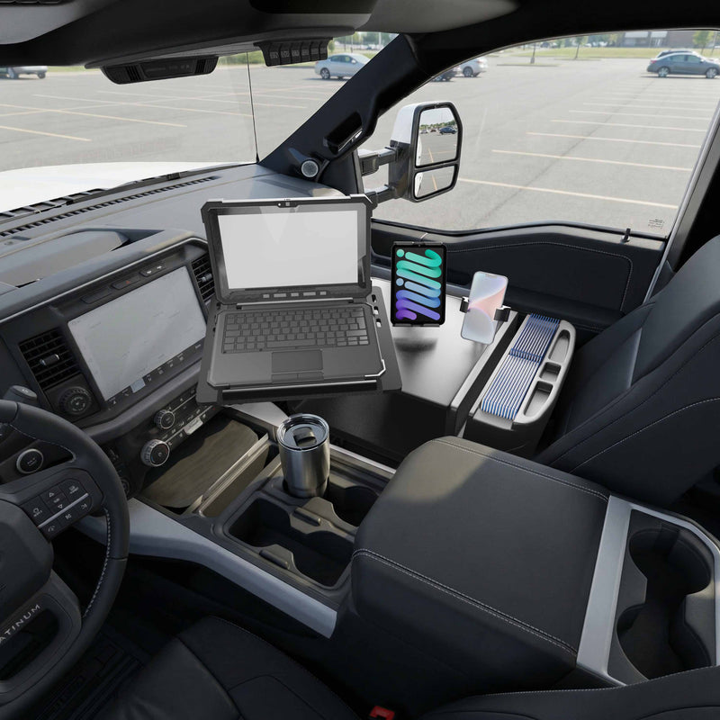 The interior of a vehicle displays an AutoExec Reach Front Seat Car Desk with Slim-Grip® Ultra Pro and RoadVise® Ultra Pro Drill-Base Mount positioned between the driver and passenger seats, neatly organizing a laptop, tablet, notepad, and coffee mug. The dedicated Slim-Grip® Ultra Pro mount securely holds your tablet on the desk, while the parking lot is visible through the windows.