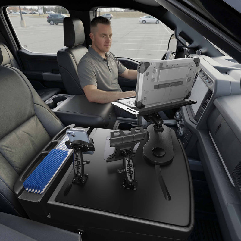 A person sits in the driver’s seat of a parked vehicle, working on an AutoExec Reach Front Seat Car Desk with Slim-Grip® Ultra Pro and RoadVise® Ultra Pro Drill-Base Mount. Additional device mounts, a tablet holder, and a blue file folder are also visible on the front center console.
