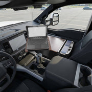 An Arkon Mounts AutoExec Reach Front Seat Car Desk holds an open rugged laptop, transforming the front passenger seat of a parked truck into a practical mobile workspace. The swivel workstation, truck dashboard, travel mug in the cup holder, and parking lot outside are all visible.