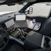 An AutoExec Reach Front Seat Car Desk with RoadVise® Ultra Pro Drill-Base Mount is set up in a truck’s front seat, transforming it into a fully functional mobile workstation. A smartphone rests securely in the RoadVise® holder, while a travel mug and other essentials fit neatly into built-in cup holders. Through the window, an empty parking lot can be seen.