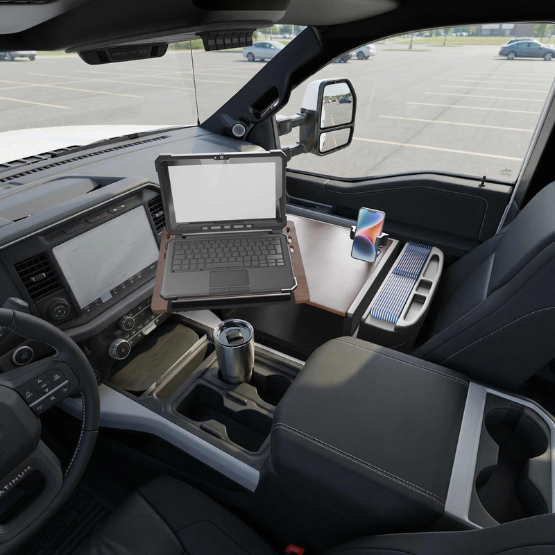 An AutoExec Reach Front Seat Car Desk with RoadVise® Ultra Pro Drill-Base Mount is set up in a truck’s front seat, transforming it into a fully functional mobile workstation. A smartphone rests securely in the RoadVise® holder, while a travel mug and other essentials fit neatly into built-in cup holders. Through the window, an empty parking lot can be seen.