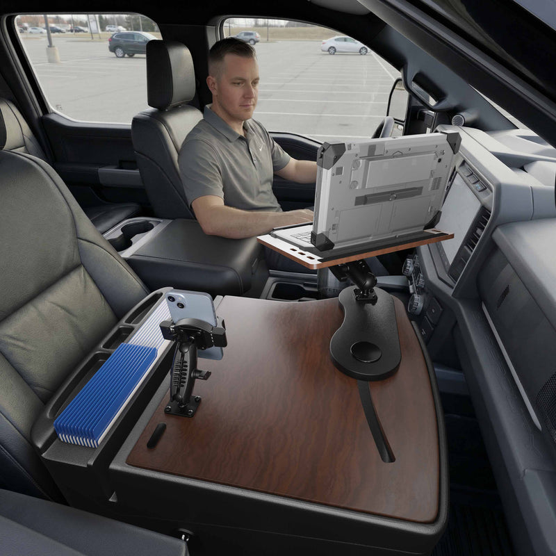 A man sits in the driver’s seat of a parked vehicle, working on his laptop mounted on an AutoExec Reach Front Seat Car Desk with RoadVise® Ultra Pro Drill-Base Mount, which is attached to the center console. The mobile workstation also features a phone holder and other office supplies.