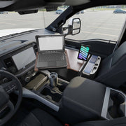 An AutoExec Reach Front Seat Car Desk with Slim-Grip® Ultra Pro Drill-Base Mount by Arkon Mounts is set up on the front passenger seat of a parked vehicle, with a laptop resting on the desk and a tablet secured in the mount, creating an efficient mobile workspace. Nearby, a travel mug completes the setup, while an empty parking lot can be seen through the window.