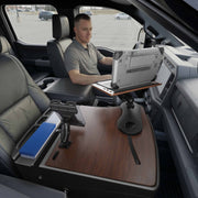 A man sits in the driver’s seat of a parked truck, working on his laptop which is placed on an AutoExec Reach Front Seat Car Desk with Slim-Grip® Ultra Pro Drill-Base Mount by Arkon Mounts. The front passenger area is set up as a mobile workspace, featuring practical additions such as a tablet holder for enhanced convenience.