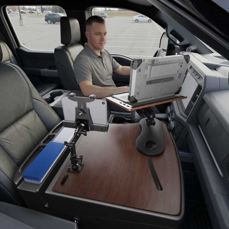 A man sits in the driver’s seat of a parked vehicle, working on a laptop placed on an AutoExec Reach Front Seat Car Desk equipped with a LockVise™ Locking Tablet Multi-Angle Mount, which securely holds another device next to a box of blue-striped index cards.