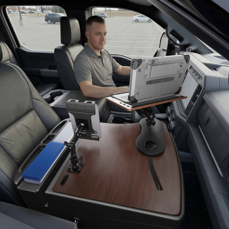 A man sits in the driver’s seat of a parked vehicle, working on his laptop at the AutoExec Reach Front Seat Car Desk by Arkon Mounts, which is installed across the center console. The desk features a Slim-Grip® Ultra Pro Multi-Angle Mount for a tablet and includes a blue file organizer, creating an efficient mobile workstation.