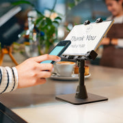 A person holds a phone near a tablet on the LockVise™ POS Locking Tablet Stand at a counter, making a contactless payment. The tablet screen shows “Thank You” and “PAID $11.00,” with a coffee cup and plant in the background.
