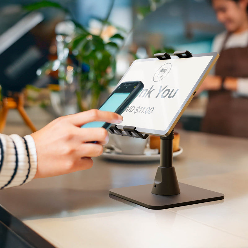 A person pays at a counter by holding their smartphone near a Slim-Grip® Ultra Pro POS Tablet Stand, which displays “Thank You” and a payment amount of $11.00 on its digital tablet screen. The background features a cafe setting with softly blurred elements.