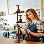 A woman wearing a plaid shirt and apron stands behind a counter, using a LockVise™ POS Locking Tablet Mount with Triple Tablet Holders that securely supports three tablet devices, with a bar setup visible in the background.