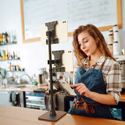 A woman in a plaid shirt and apron uses a Slim-Grip® Ultra Pro POS Tablet Mount with Triple Holders to manage three devices in a modern café setting. Shelves, cups, and menu boards are visible in the background, highlighting how this innovative retail POS tablet mount streamlines order processing and customer service.