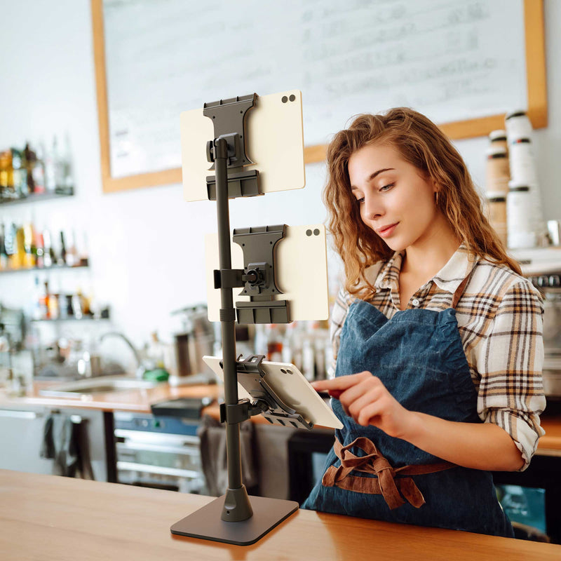 A woman in a plaid shirt and apron uses a Slim-Grip® Ultra Pro POS Tablet Mount with Triple Holders to manage three devices in a modern café setting. Shelves, cups, and menu boards are visible in the background, highlighting how this innovative retail POS tablet mount streamlines order processing and customer service.