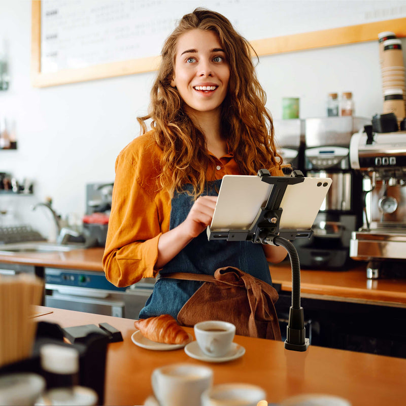 A smiling barista with long curly hair stands behind a coffee shop counter, using a touchscreen device secured in the LockVise™ POS Locking Tablet Holder with 12” Gooseneck Clamp Mount. A croissant and a cup of coffee sit on the counter in front of her.