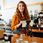 A smiling woman in a café stands behind the counter with a tablet mounted on the Slim-Grip® Ultra Pro POS Tablet Mount with 12” Gooseneck Clamp, a coffee cup and croissant in front of her, and a coffee machine in the background.