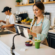 A smiling cafe worker uses a touchscreen register mounted on the Slim-Grip® Ultra Pro POS Tablet Mount with 12” Gooseneck Clamp next to a smoothie and acai bowl, while another employee prepares food in the background.