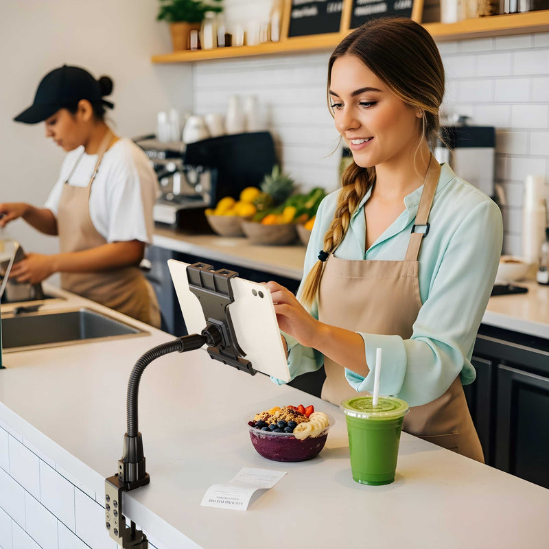 A smiling cafe worker uses a touchscreen register mounted on the Slim-Grip® Ultra Pro POS Tablet Mount with 12” Gooseneck Clamp next to a smoothie and acai bowl, while another employee prepares food in the background.
