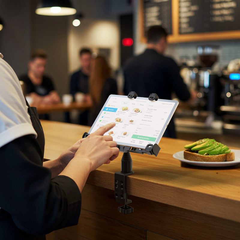 A person uses the LockVise™ Locking POS Tablet Mount with Clamp Mount to securely place a food order on a mounted tablet at a café counter. In the background, people gather near the barista area, and a plate of avocado toast rests on the counter.
