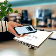 A person holds a smartphone near a digital payment terminal displaying “Thank You PAID $11.00,” indicating a contactless payment. The Slim-Grip® Ultra Pro POS Drill-Base Tablet Mount securely holds the device in place, ensuring smooth and efficient transactions.