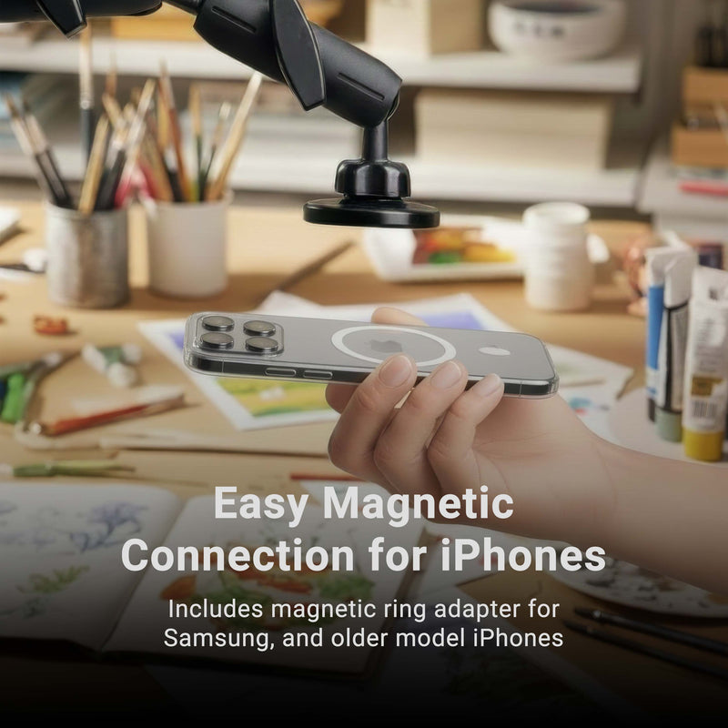 A hand holds an iPhone near the Remarkable Creator™ Pro+Plus Clamp Mount - MagSafe Compatible above a cluttered desk with art supplies. Text reads: Easy magnetic connection for iPhones. Includes magnetic ring adapter for Samsung, older model iPhones, and is compatible with most creator clamp mounts and overhead phone mounts.
