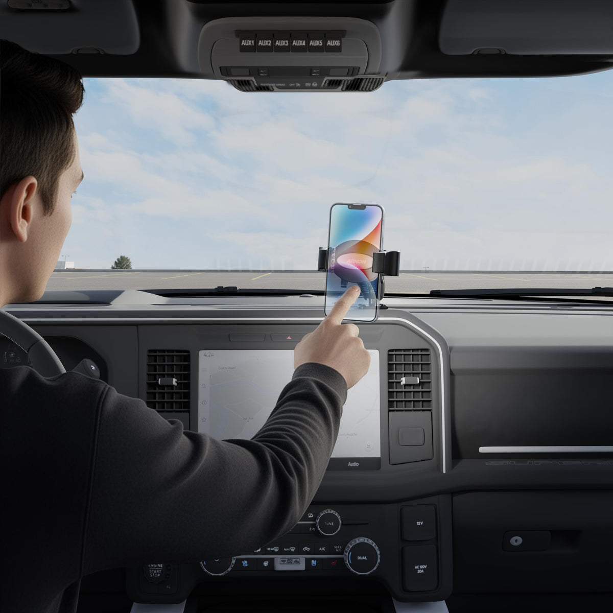 A person sitting in a car uses their right hand to touch a smartphone secured in a RoadVise® Ultra Pro Phone Holder 5” AMPS Drill-Base Mount on the dashboard, with the car’s interior and the sky through the windshield visible.