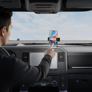 A person sitting in a car uses their right hand to touch a smartphone secured in a RoadVise® Ultra Pro Phone Holder 5” AMPS Drill-Base Mount on the dashboard, with the car’s interior and the sky through the windshield visible.
