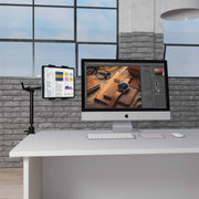 A modern office desk features an iMac displaying photo editing software, a wireless keyboard and mouse, and a tablet securely held by the Arkon Mounts Slim-Grip® Tablet Holder with Clamp Mount and 22″ Arm, all set against large windows and exposed brick walls in the background.