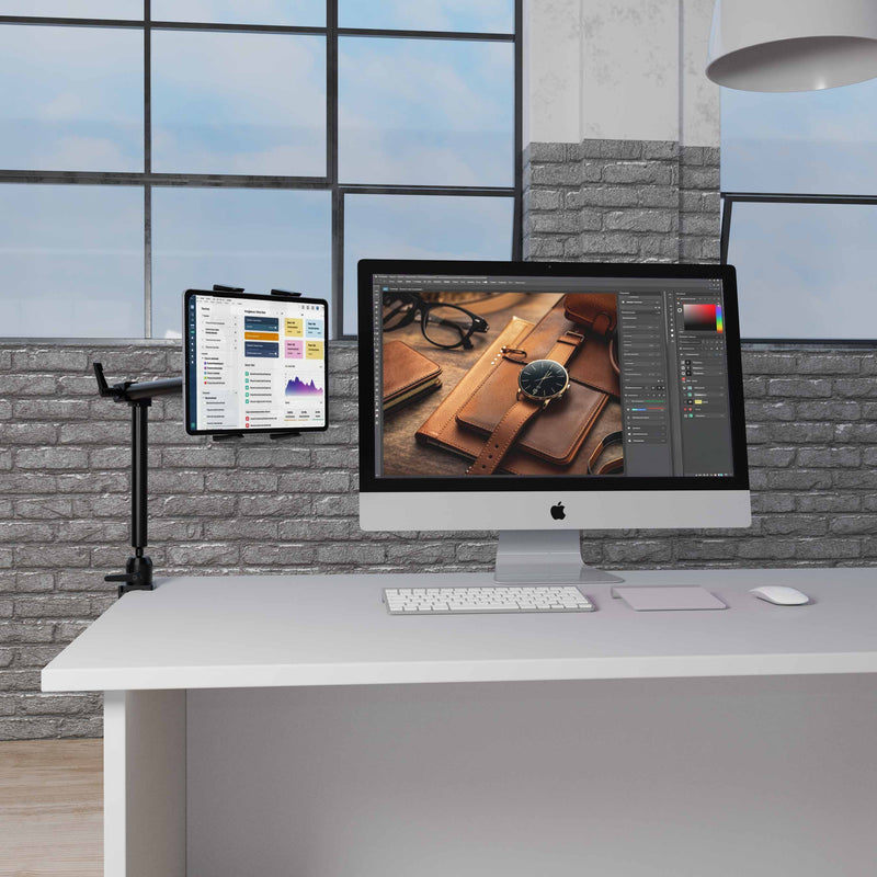 A modern office desk features an iMac displaying photo editing software, a wireless keyboard and mouse, and a tablet securely held by the Arkon Mounts Slim-Grip® Tablet Holder with Clamp Mount and 22″ Arm, all set against large windows and exposed brick walls in the background.