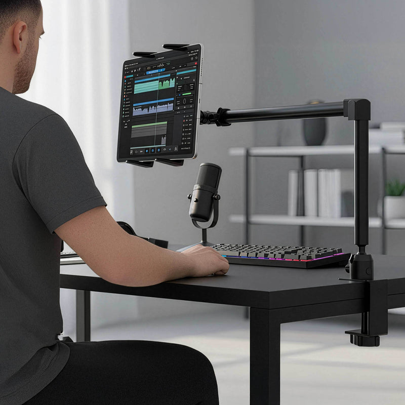 A person sits at a desk using a tablet held securely by the Arkon Mounts Slim-Grip® Tablet Holder with Clamp Mount and 22″ Arm. In front of them are a microphone and a colorful keyboard, while audio editing software is visible on the tablet screen. The heavy-duty clamp mount ensures the setup stays stable during use.
