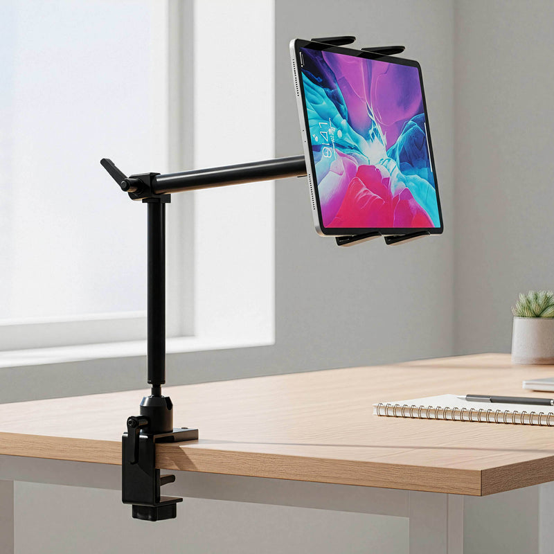 A tablet is securely held by the Arkon Mounts Slim-Grip® Tablet Holder with Clamp Mount and 22″ Arm, which is attached to the edge of a wooden desk. The workspace also features a spiral notebook, pen, and small potted plant near a bright window.