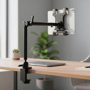 An Arkon Mounts Slim-Grip® Tablet Holder with Clamp Mount and a 22″ adjustable black arm is secured to a wooden desk, holding a tablet above a closed laptop. In the background, a potted plant is softly blurred.