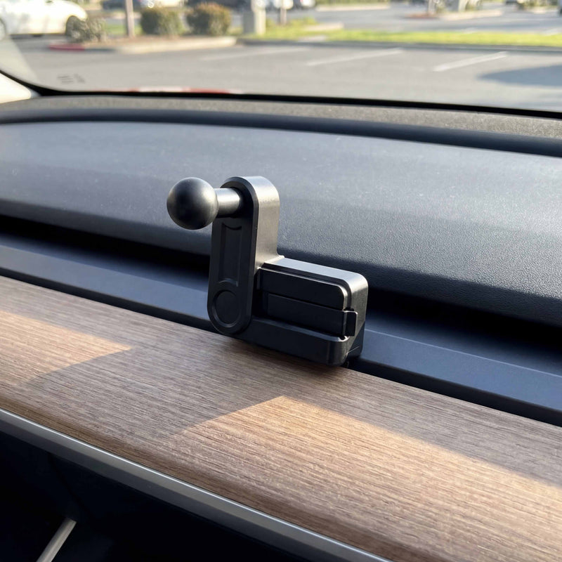 A Vent Mount for Tesla Model 3 (2017-2022) and Model Y (2020-2023) made of black plastic with a ball joint is attached to the car’s wood trim dashboard in sunlight, with a parking lot and trees visible through the windshield.