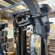 A close-up of the Arkon Mounts XMS™ Heavy Duty Forklift Mount with AMPS Adapter Plate attached to a forklift frame, with a mirror and stacked boxes visible in the background. The mount is secured with bolts and adjustment knobs.