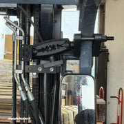 Close-up of a forklift equipped with the Arkon Mounts XMS™ Heavy Duty Forklift Mount with AMPS Adapter Plate, attached near the mirror. Boxes and pallets can be seen in the background, while the focus remains on the mounting equipment and hydraulic hoses.