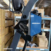 A tablet is mounted on a forklift using the XMS™ Heavy Duty Forklift Mount with LockVise™ Locking Tablet Holder, as part of a reliable warehouse tablet mounting system, in a warehouse filled with cardboard boxes on metal shelves.