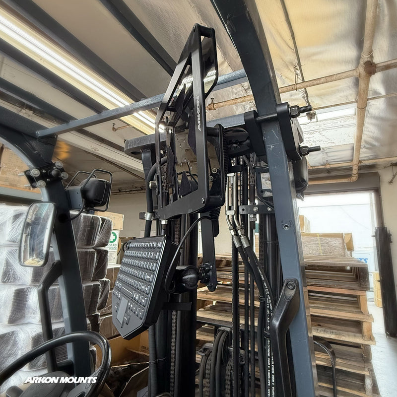 A forklift equipped with an Arkon Mounts XMS™ Heavy Duty Forklift Mount featuring a Dual Utility Mounting Bracket for securely attaching a keyboard and screen near the controls, utilizing VESA AMPS compatibility, positioned inside a warehouse with stacked pallets and boxes in the background.