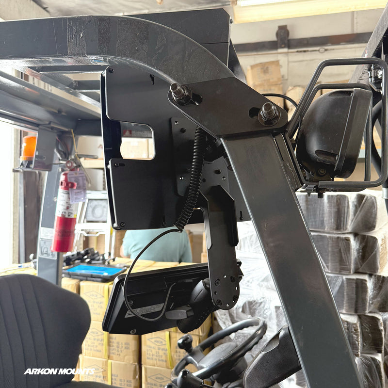 Close-up of a forklift interior featuring the Arkon Mounts XMS™ Heavy Duty Forklift Mount with Dual Utility Mounting Bracket, VESA AMPS compatible, securely installed next to a rearview mirror and fire extinguisher. Stacks of boxes are visible in the background, with the Arkon Mounts logo in the lower left.