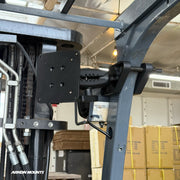 The XMS™ Heavy Duty Forklift Mount with VESA Adapter Plate is securely attached to the metal frame of a forklift, with cardboard boxes visible in the background. The scene takes place in a warehouse or storage facility typical of material handling environments.