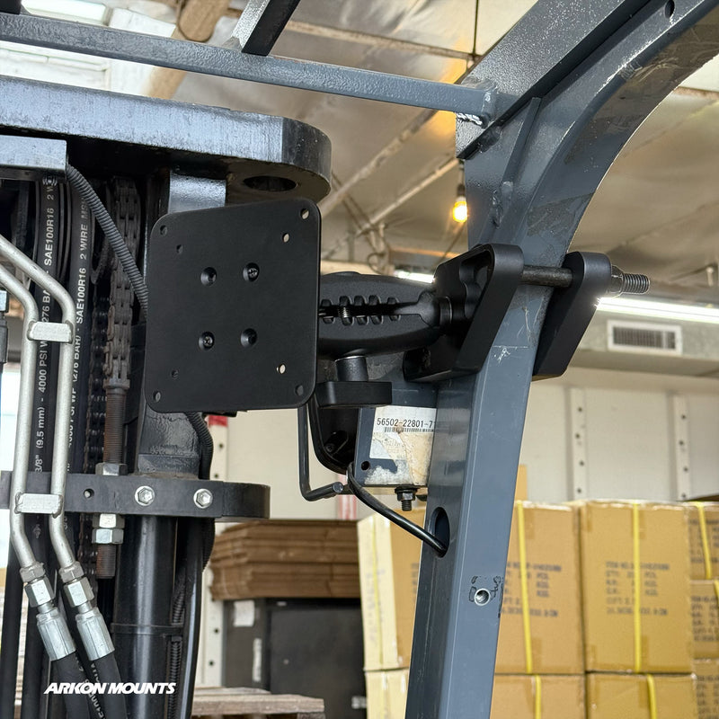The XMS™ Heavy Duty Forklift Mount with VESA Adapter Plate is securely attached to the metal frame of a forklift, with cardboard boxes visible in the background. The scene takes place in a warehouse or storage facility typical of material handling environments.