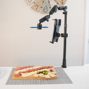 The Remarkable Creator™ Pro+Plus with Clamp Mount holds a smartphone above a table, capturing a top-down shot of a wooden board with meats, cheeses, crackers, and olives. A large wall clock appears in the background.