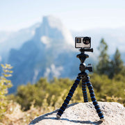 A Flexible 11 Tripod for GoPro HERO Action Cameras holds a small action camera on a rock, with mountains and trees softly blurred in the background under a clear sky.