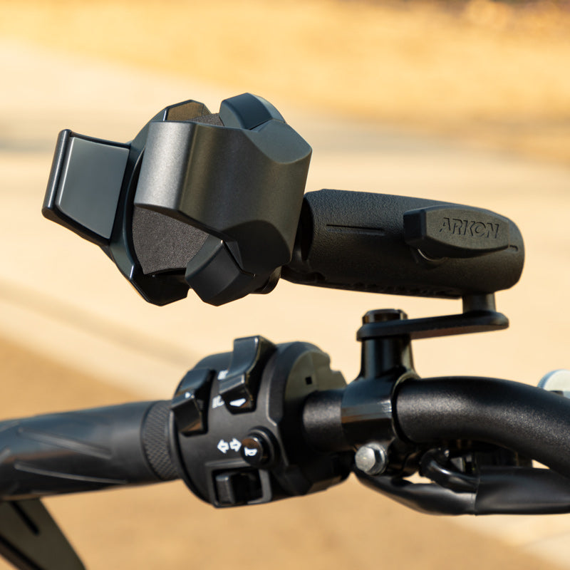 Close-up of the RoadVise® Ultra Motorcycle Handlebar Mirror Pinch Bolt Stem Phone and Tablet Mount, attached beside bike controls and textured grips on a handlebar, set against a blurred outdoor background.