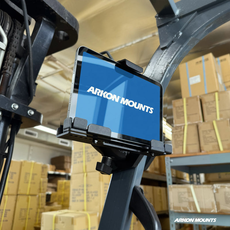 A tablet secured in the XMS™ Heavy Duty Forklift Mount with LockVise™ Metal XL Locking Tablet Holder from Arkon Mounts is attached to warehouse equipment, surrounded by shelves stacked with cardboard boxes. The tablet displays the Arkon Mounts logo on a blue screen.